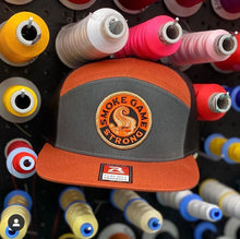 Load image into Gallery viewer, Richardson&#39;s 7 panel hat in fire glow orange and grey with Smoke Game Strong logo in orange, khaki and black. Hat is sitting among spools of thread.
