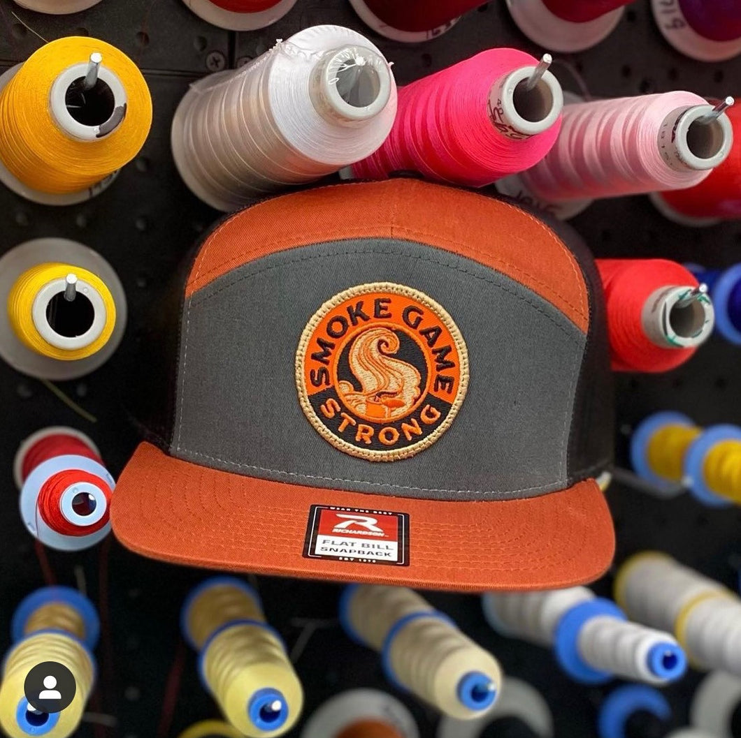 Richardson's 7 panel hat in fire glow orange and grey with Smoke Game Strong logo in orange, khaki and black. Hat is sitting among spools of thread.